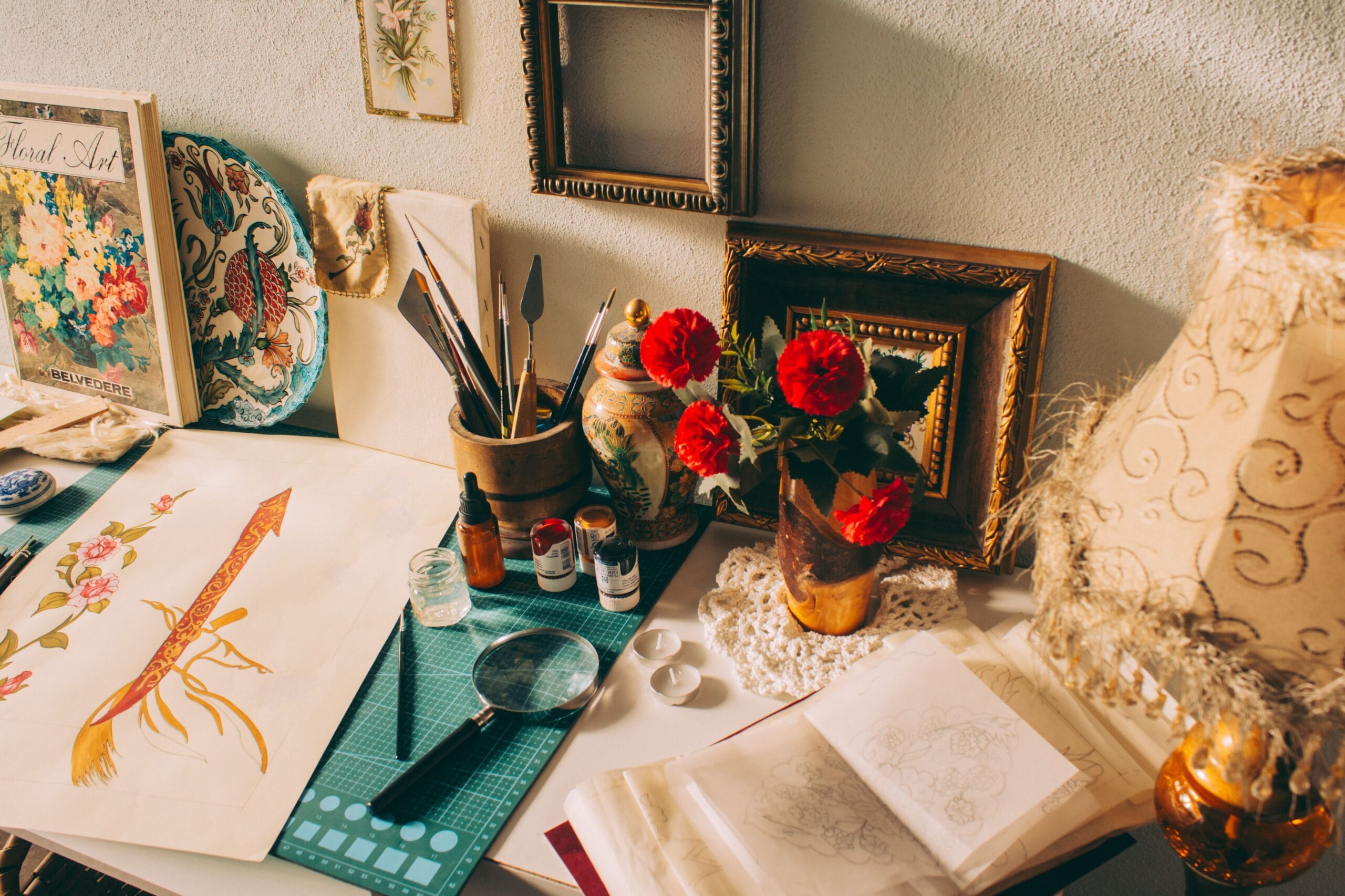 A sunlit worktable with brushes, inks, floral studies in progress and a jar of red carnations