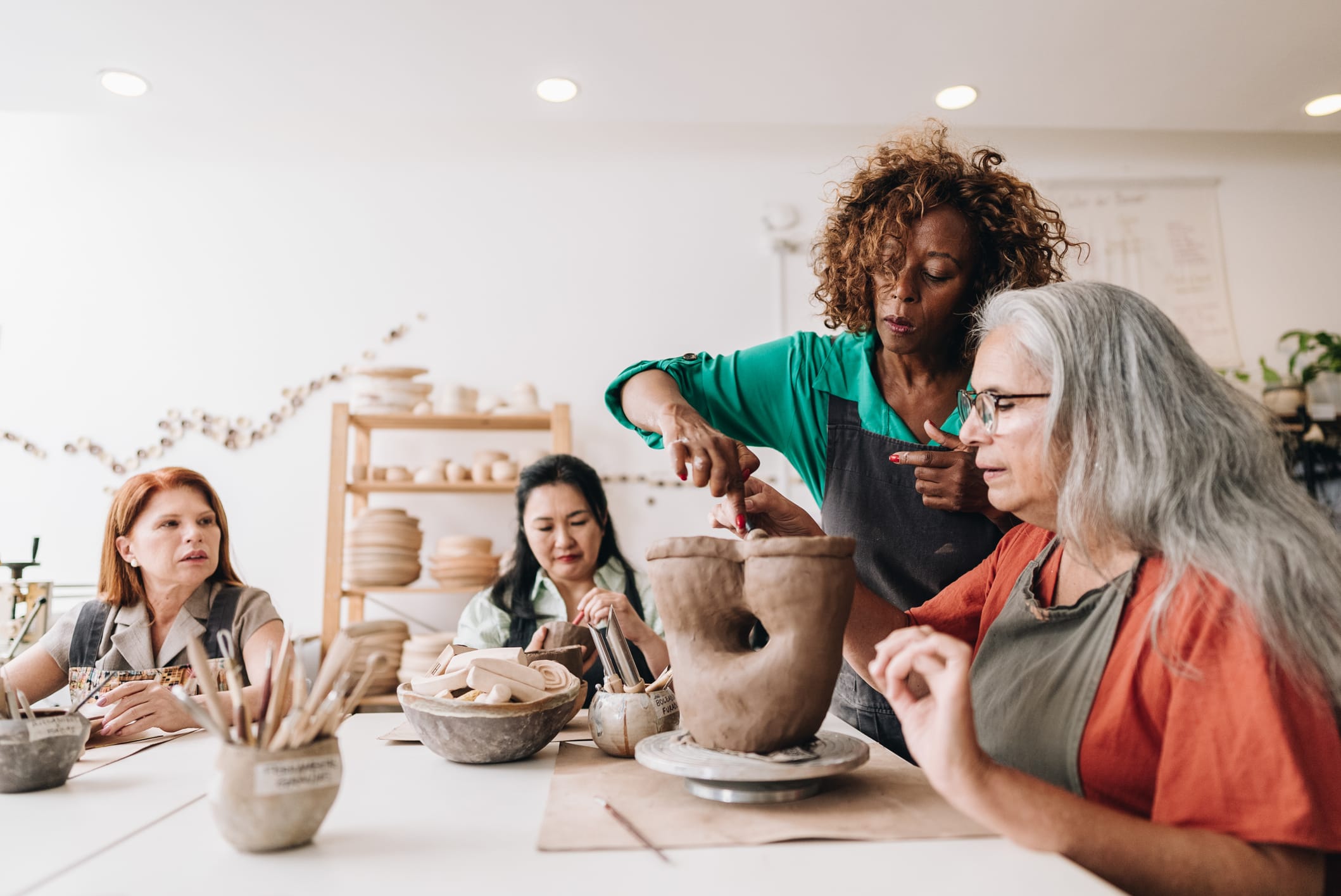 A teacher leaning in to guide a hand-built pot while the rest of a small pottery class works around the table