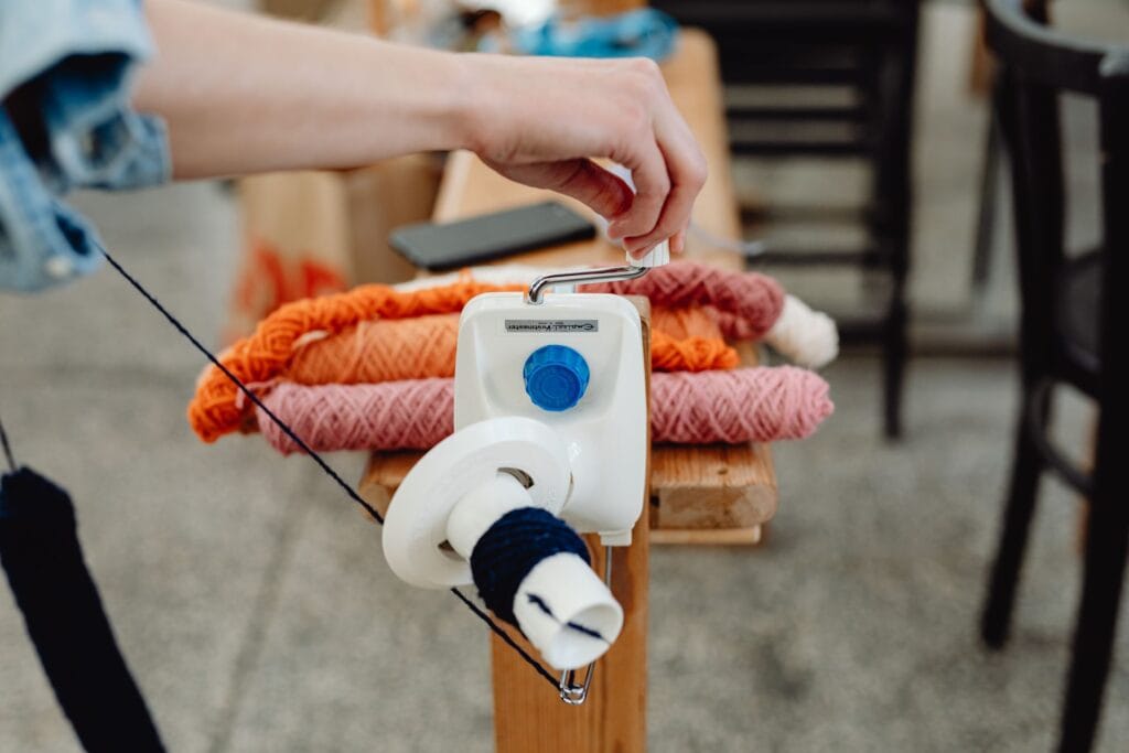 A hand turning a manual yarn winder beside skeins of colorful wool