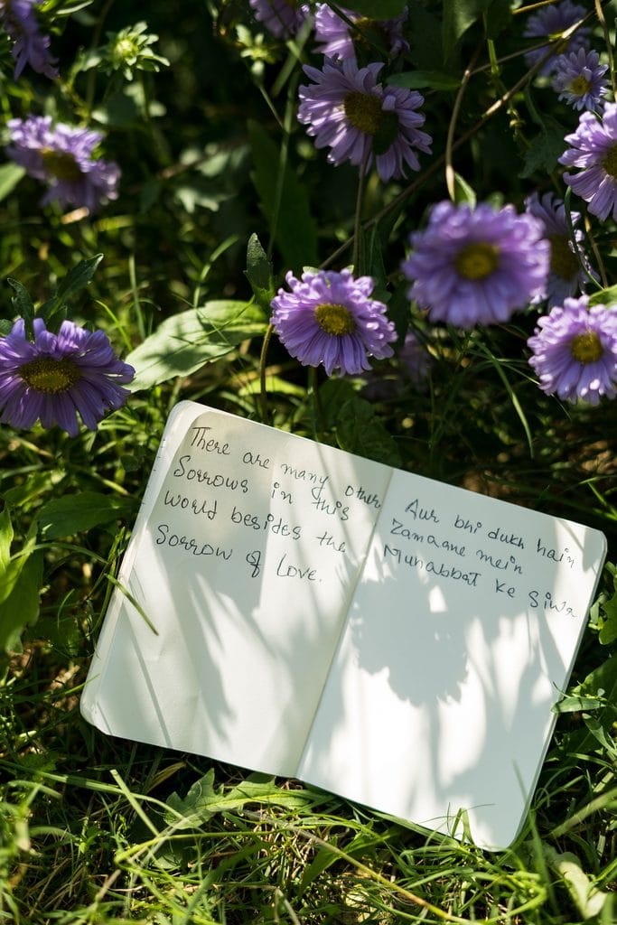 A handwritten poem rests open on a white notebook among purple flowers and green grass, with sunlight and leafy shadows falling across the pages.